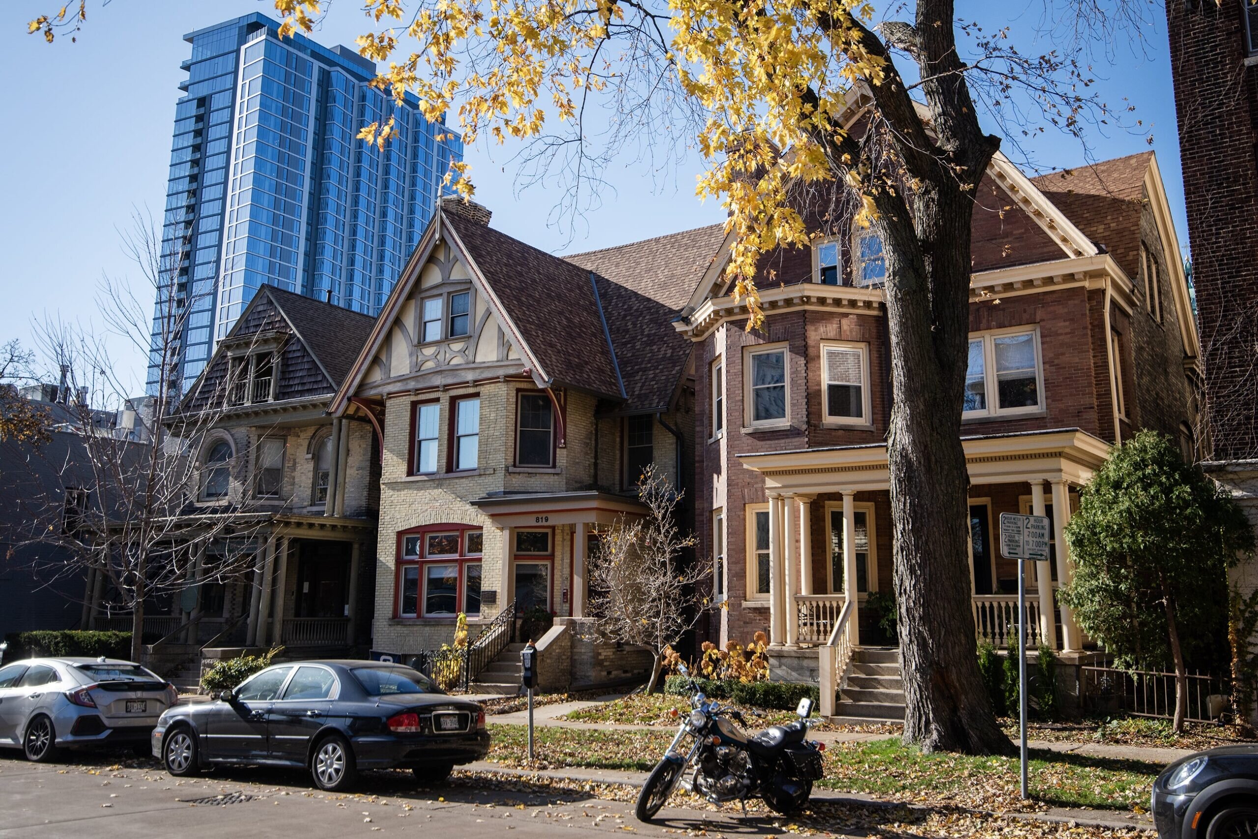 A row of charming vintage homes with unique architectural designs, including gabled roofs, detailed brickwork, and spacious porches. Autumn leaves and trees frame the scene, with a modern glass high-rise building towering in the background, creating a blend of historic and contemporary urban landscape.