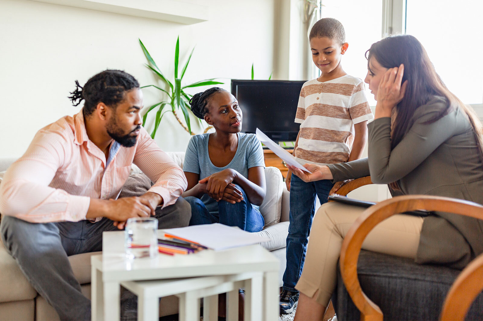A family of four, including a man, a woman, and two children, is sitting on a couch in their living room. The woman is attentively listening as a real estate professional reviews documents with them, highlighting a consultative and family-oriented real estate experience.