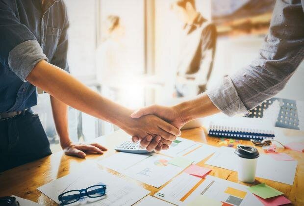 Two individuals shaking hands over a desk, symbolizing a successful business agreement or partnership, with documents and office items in the background.
