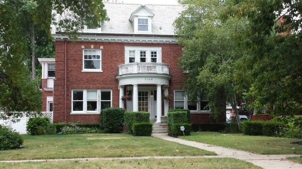 A large, red-brick, colonial-style house with white-trimmed windows and an elegant rounded entryway supported by white columns. The home is surrounded by lush greenery and mature trees.