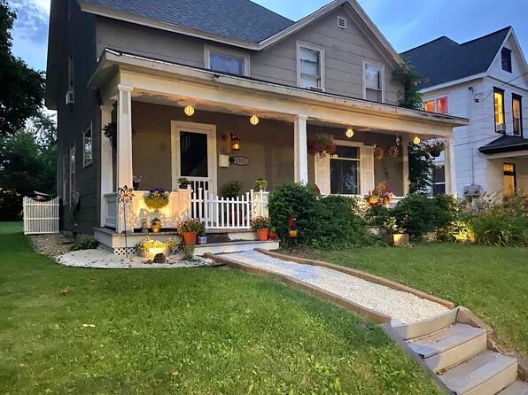 A cozy, well-maintained house with a charming front porch decorated with hanging lights, potted plants, and flower baskets. A white picket fence and a pathway lined with subtle lighting enhance the home's welcoming ambiance during dusk.