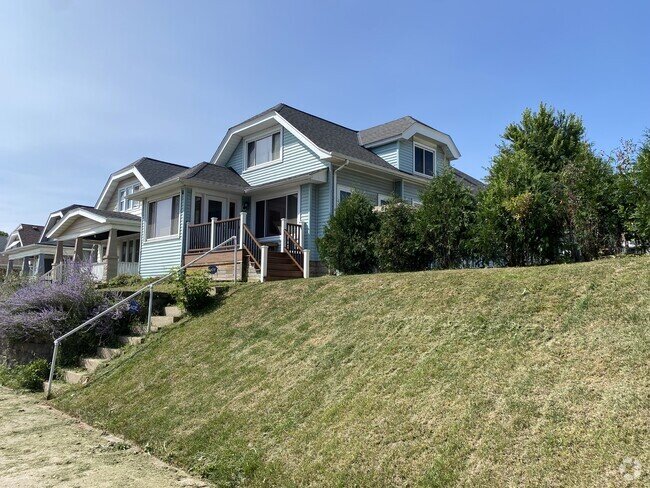 A light blue, two-story home sitting on a slightly elevated lawn. The house features a charming porch with wooden stairs and a small front yard, and is flanked by similar houses in a suburban neighborhood.