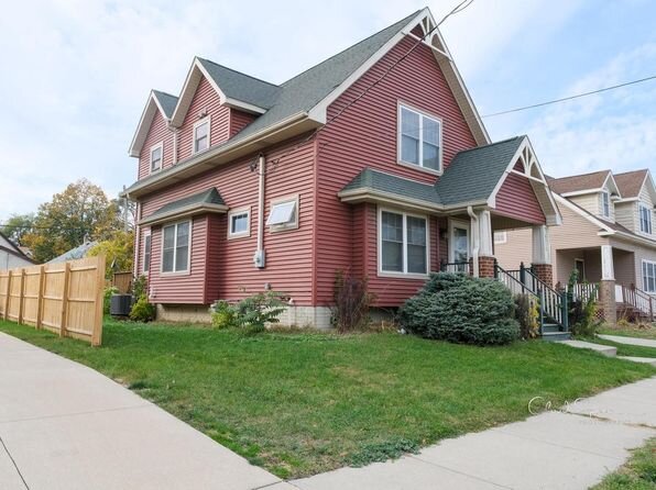 A charming red two-story house featuring white trim, a steeply pitched roof, and a well-maintained yard. The home is bordered by a wooden fence along a neatly paved sidewalk, adding to the neighborhood’s inviting atmosphere.