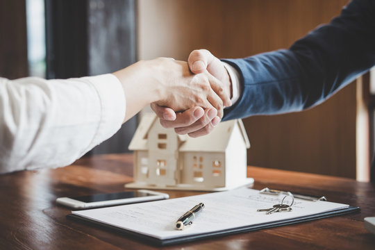 A close-up view of two people shaking hands over a real estate contract, with small model houses on the table, symbolizing an agreement on a house sale or purchase.