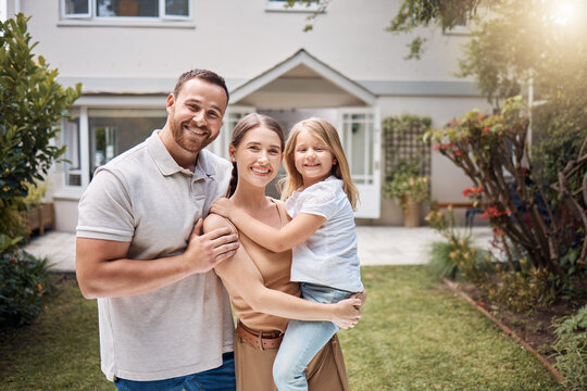 A happy family of three posing outside their new home. The father stands to the left, holding his arm around his smiling wife, who is carrying their young daughter. The family stands in a lush, well-kept garden, with the entrance of a beautiful house visible in the background."