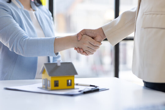 Close-up of a handshake between two individuals, with a small model house placed on a table beside them, symbolizing the closing of a real estate deal.