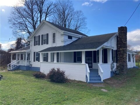A classic, two-story white farmhouse with black shutters and a large, wraparound porch. The house has an extended front area with multiple windows and a tall stone chimney on one side, all set on a grassy lawn under a partly cloudy sky.