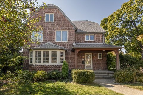 A charming two-story brick house with a quaint front garden. The home features a bay window, a covered front porch, and mature trees surrounding the property, creating a warm and inviting atmosphere.