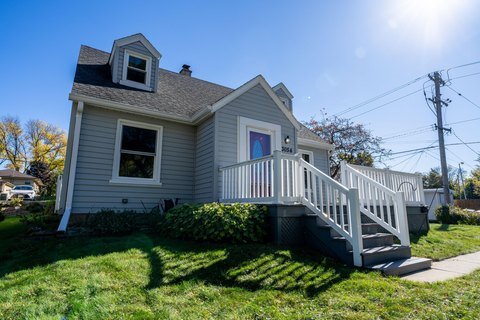 A quaint gray cottage-style home with white trim, featuring a small porch with stairs leading up to the entrance. The house is surrounded by green grass and receives ample sunlight, creating a cozy and inviting feel.