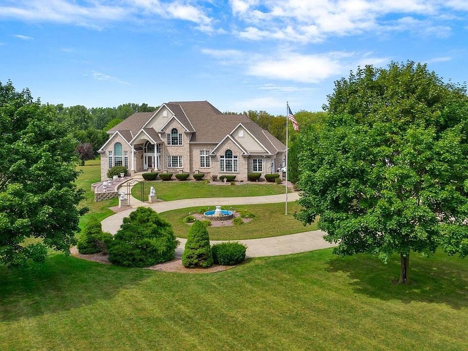 A spacious luxury estate with a grand entrance, circular driveway, and beautifully landscaped front yard. The house is set on a large green lawn with tall trees surrounding the property. A fountain is centered in front of the house, adding an elegant touch to the expansive yard. The exterior of the house is beige with large windows and a complex roof