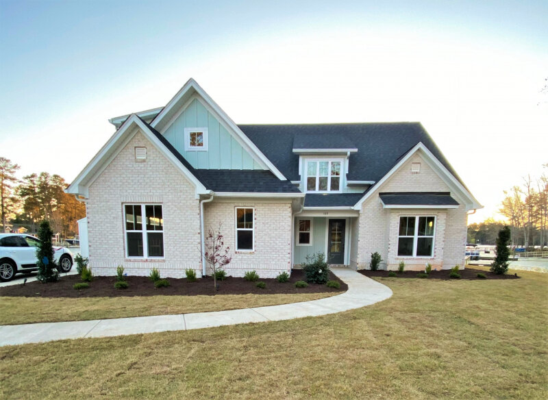 A modern single-story house with white brick walls and dark gray roof shingles. The front facade features large windows and a centered entryway. A landscaped pathway leads to the entrance, with young trees and manicured shrubs accentuating the clean, minimalist yard.
