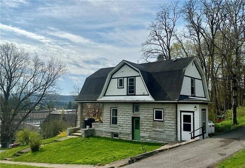 Unique stone and wood house with a gambrel roof and green doors, set on a slight hill. The home has a rustic charm with a mix of traditional architecture and a modern twist, surrounded by a modest lawn and overlooking a scenic view of the town below.