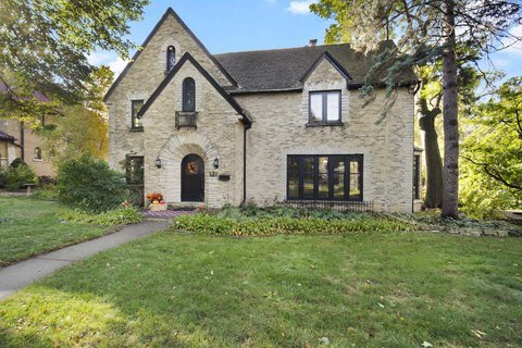 A stately stone Tudor house with a steeply pitched roof and arched entryway, set on a spacious, tree-lined lot with lush greenery, showcasing timeless architecture and a classic exterior.