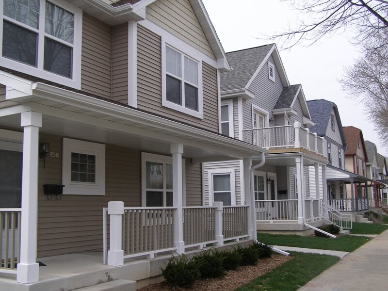 A row of modern, suburban-style townhomes with beige siding and white trim. Each home features a front porch with white railings, and a small patch of greenery borders the sidewalk. The townhomes are uniformly designed, showcasing a cohesive and tidy residential community.