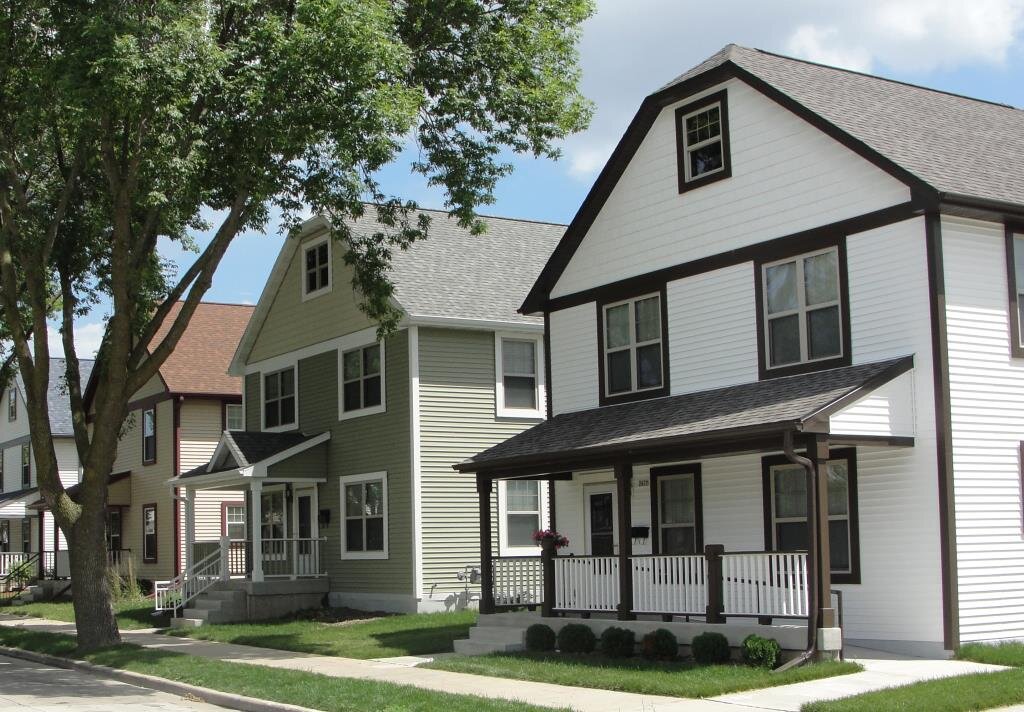 A row of three houses with modern, suburban architecture, featuring neutral-colored siding and white trim. The foreground house has a spacious front porch and a sloped roof, set among mature trees and a well-maintained sidewalk.