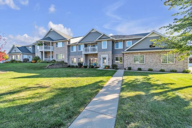 A modern two-story apartment complex with beige and gray exterior, large windows, and individual balconies. The building sits on well-maintained green lawns with a pathway leading to the entrances.