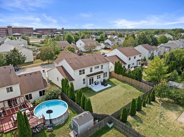 A suburban backyard with an above-ground pool, fenced-in yard, and neighboring houses. The homes are situated in a well-maintained residential area with a clear sky in the background.