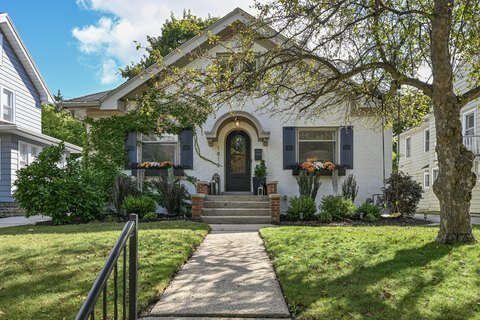 A cozy single-story home with light brick walls partially covered in ivy. The front yard is well-maintained, with a small pathway leading up to a dark arched front door adorned with seasonal flowers, adding a touch of warmth to the overall look.