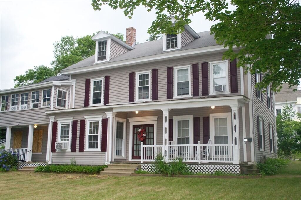A classic two-story colonial-style home with gray siding and burgundy shutters. The house features a welcoming front porch with white pillars, a well-maintained lawn, and a traditional red door accented by a seasonal wreath. The front yard is shaded by mature trees, enhancing the home's traditional charm.