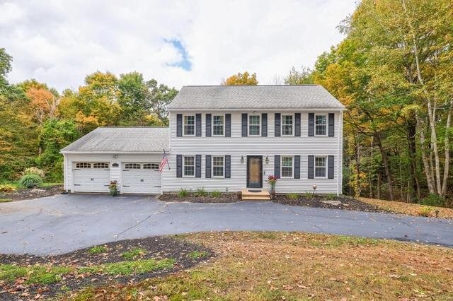 A charming two-story white colonial house with black shutters, a two-car garage, and a neatly maintained front lawn, set against a backdrop of trees with autumn foliage.