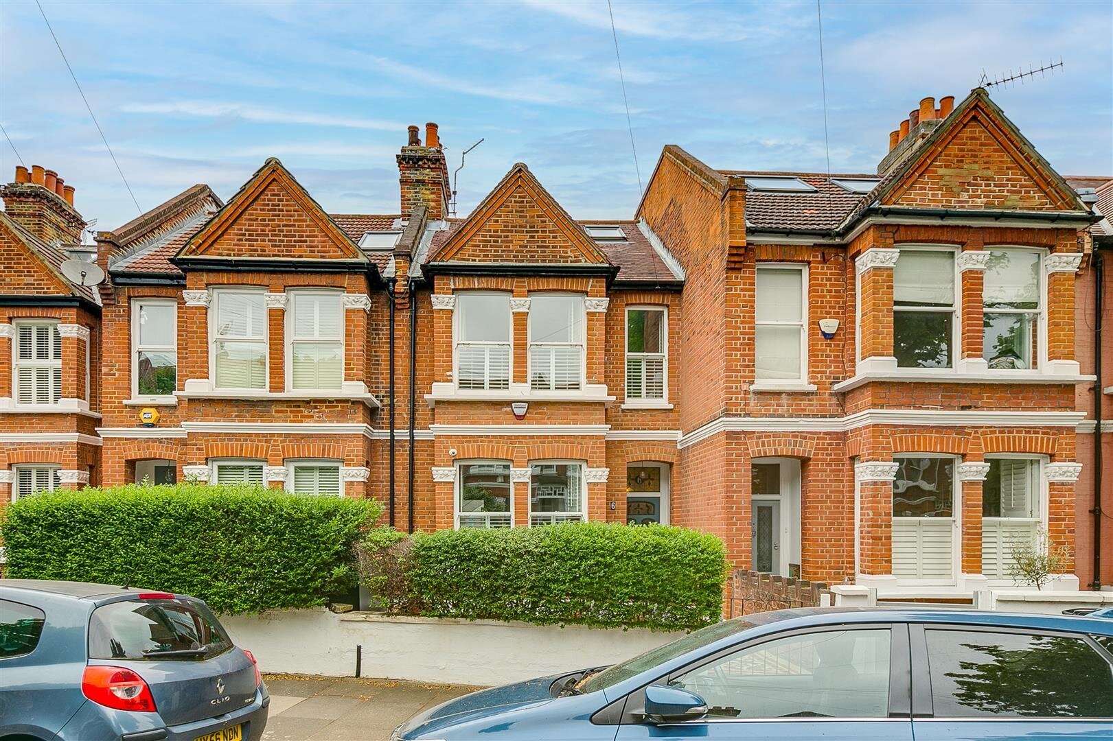 A row of classic brick townhouses with intricate white window trim and steep triangular roofs. The street view showcases neatly maintained hedges and parked cars in a quiet residential area.
