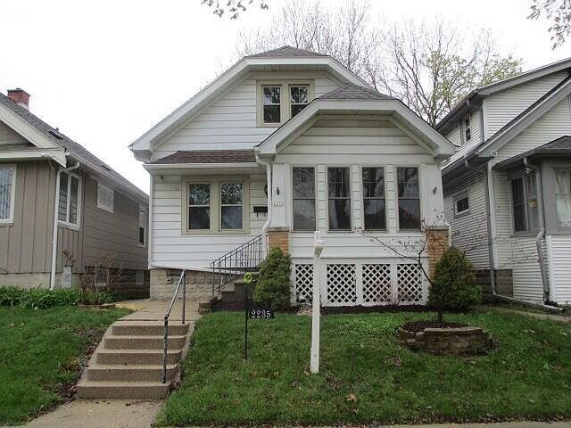 A white single-story house with an enclosed front porch, located between two neighboring homes. The house features a small front lawn, a staircase leading up to the porch, and an attached garage.