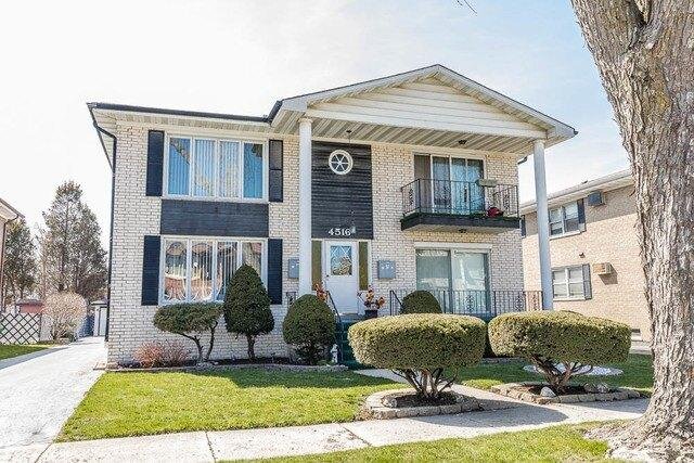 A white duplex building with black shutters and two levels. The front features a small balcony on the second floor, and well-manicured shrubs are arranged along the entry path, creating a neat and tidy front yard.