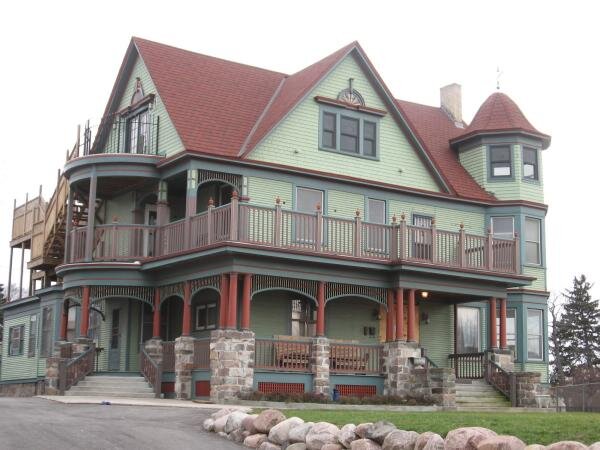 A charming Victorian-style house painted in green with red and brown accents. The house features a wrap-around balcony with ornate woodwork and a distinctive red roof, adding to its historic charm. The stone foundation adds a rustic touch to the overall aesthetic.
