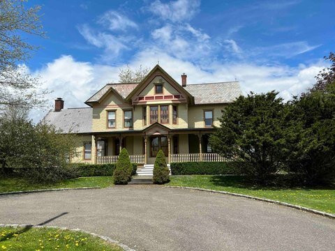 A charming Victorian-style house with a two-story design, featuring a large porch supported by decorative columns. The house is surrounded by greenery, including manicured bushes and tall trees, and set against a backdrop of a blue sky with scattered clouds.