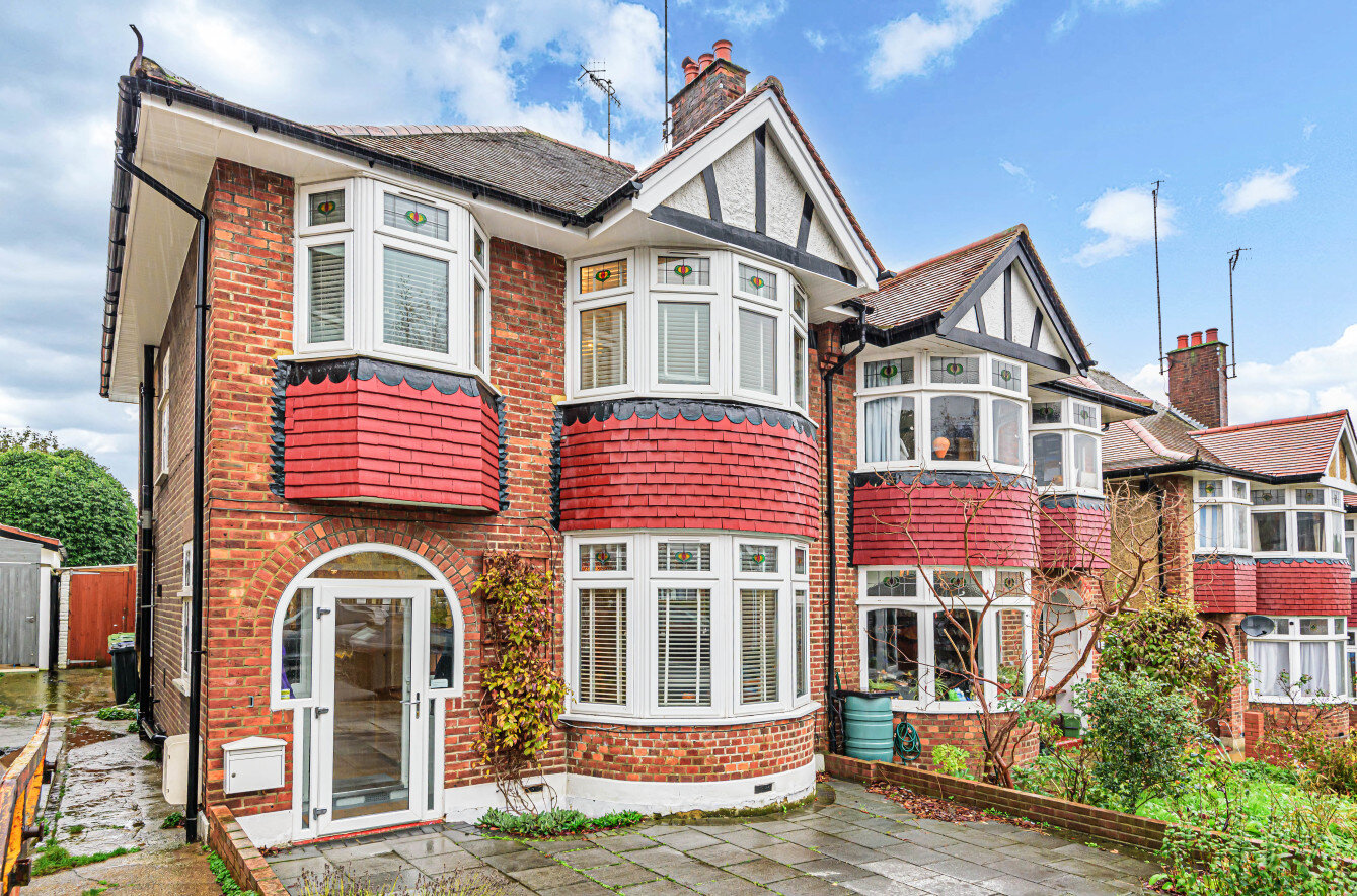 charming red-brick semi-detached house featuring large bay windows with white frames, red shingle accents, and Tudor-style detailing on the upper facade. The paved driveway leads to an arched entrance, with ivy growing on the walls and decorative greenery in the surroundings.