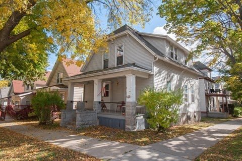 A cozy, two-story house with a gray exterior, wide front porch, and columns supporting the roof. The house is set along a tree-lined street with autumn leaves scattered on the sidewalk and grass. Bright sunlight highlights the fall foliage around the property.