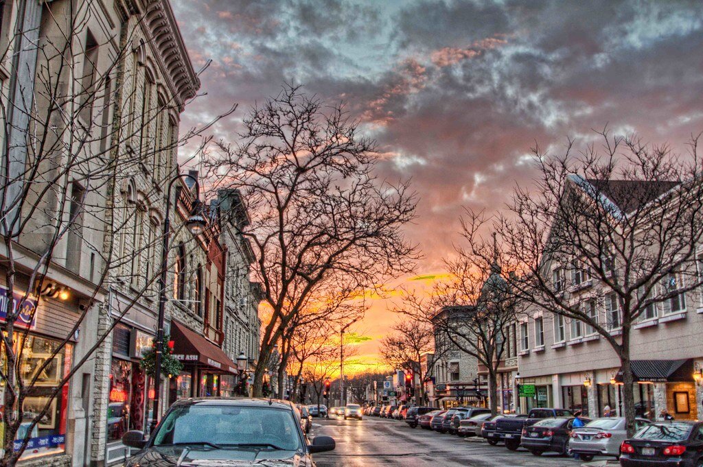 A picturesque view of a small town's main street at sunset, with historic buildings lining the street and bare trees silhouetted against a colorful sky.