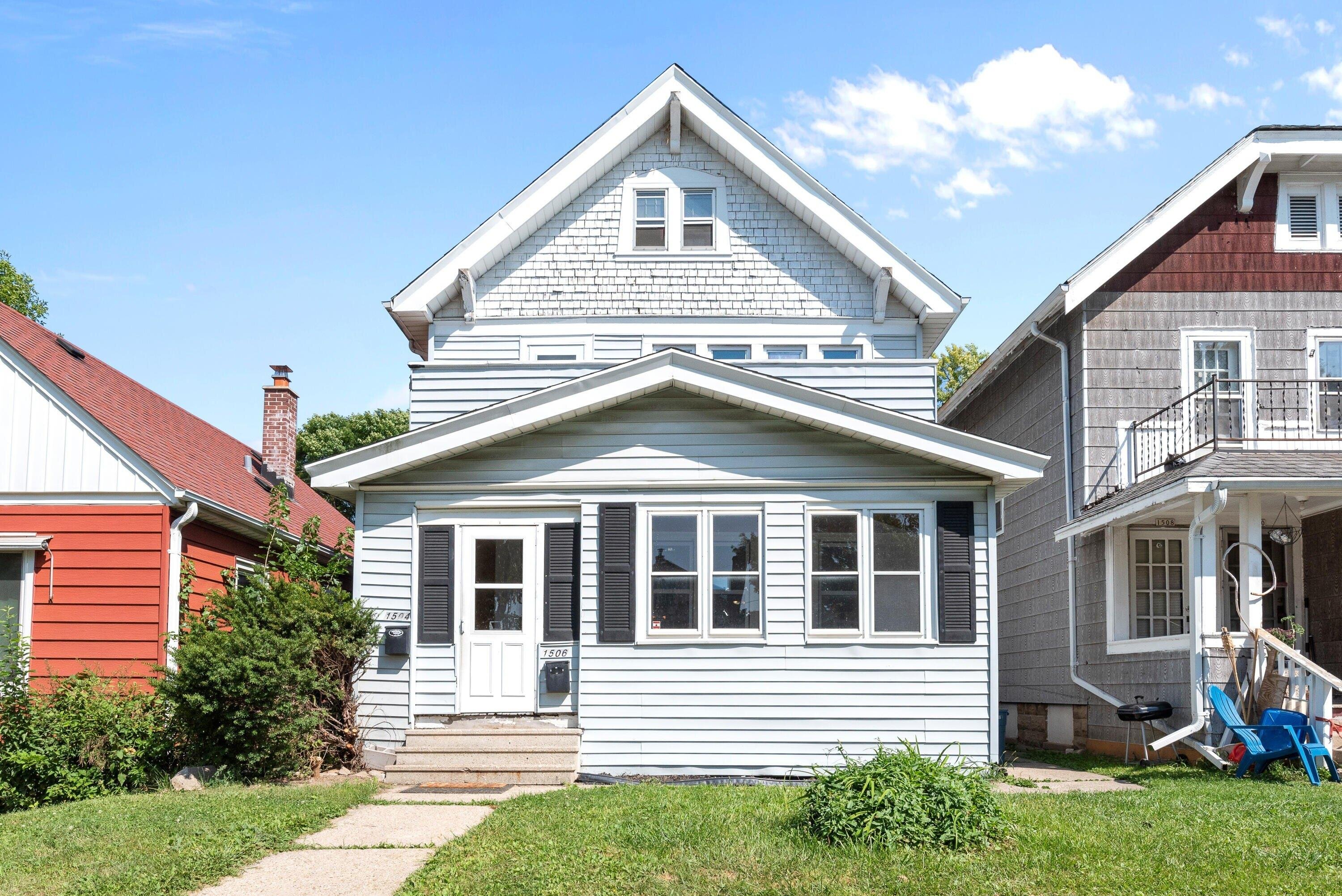 A charming two-story house with light blue siding and a gabled roof, nestled between two neighboring homes. The house has white trim around the windows and a small set of steps leading up to the front door, framed by black shutters on either side.