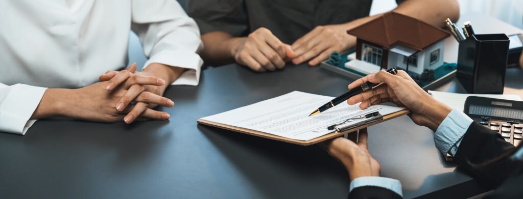 A real estate contract signing session with a real estate agent presenting a document to two clients. The table has a small house model, calculator, and pen, indicating a formal property transaction. The clients are seated with their hands folded, listening attentively to the agent's explanation.