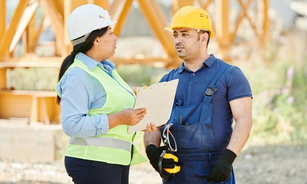 Two construction workers in hard hats and safety vests having a discussion at a construction site. One worker is holding a clipboard and pen, while the other is listening attentively, creating a scene that highlights teamwork and project management in construction.