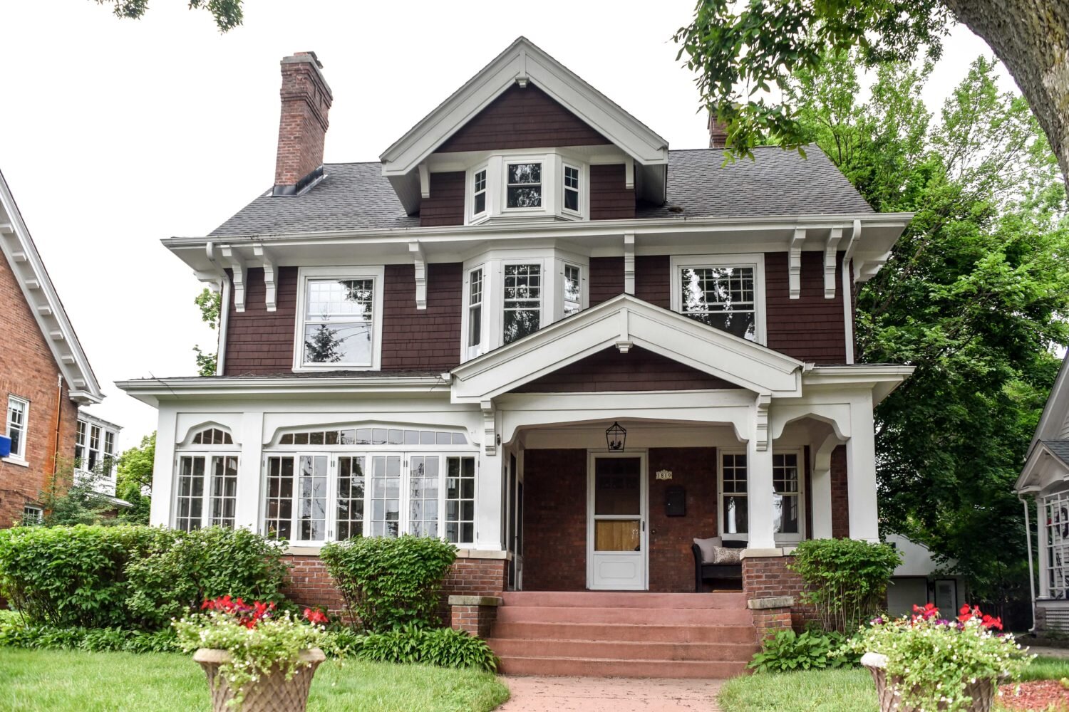 A beautiful two-story house with dark brown exterior and white trim accents. The home features large windows, a covered porch, and a prominent chimney. Lush green shrubs line the front yard, with red flowers in the foreground adding a pop of color.