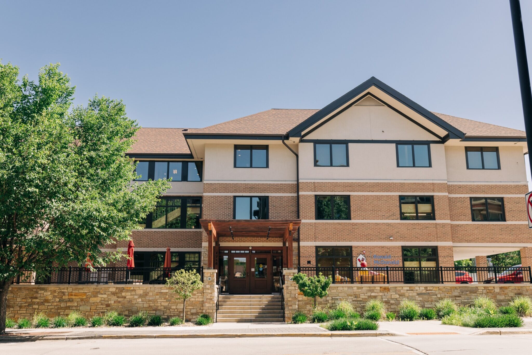 A contemporary two-story building with beige and brick accents, designed with large windows and a welcoming entrance. The building is surrounded by greenery, and a spacious patio area sits in front, offering a well-maintained outdoor space. The structure combines modern architectural elements with natural landscaping.