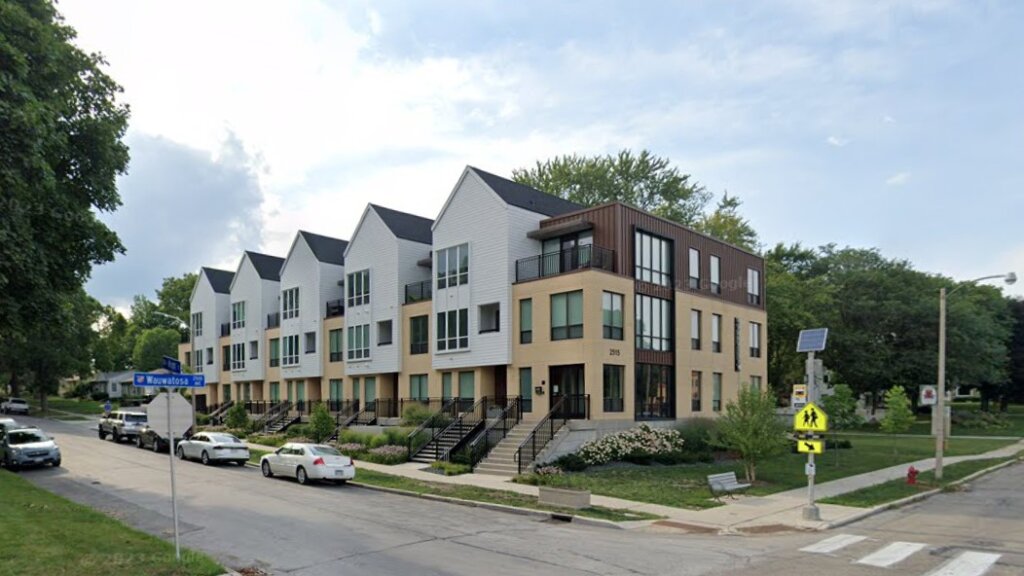 A modern row of townhouses with clean, angular designs and light-colored facades, located on a street corner. The landscaping is well-maintained, and the overall scene presents a contemporary and suburban vibe.