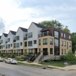 A modern row of townhouses with clean, angular designs and light-colored facades, located on a street corner. The landscaping is well-maintained, and the overall scene presents a contemporary and suburban vibe.