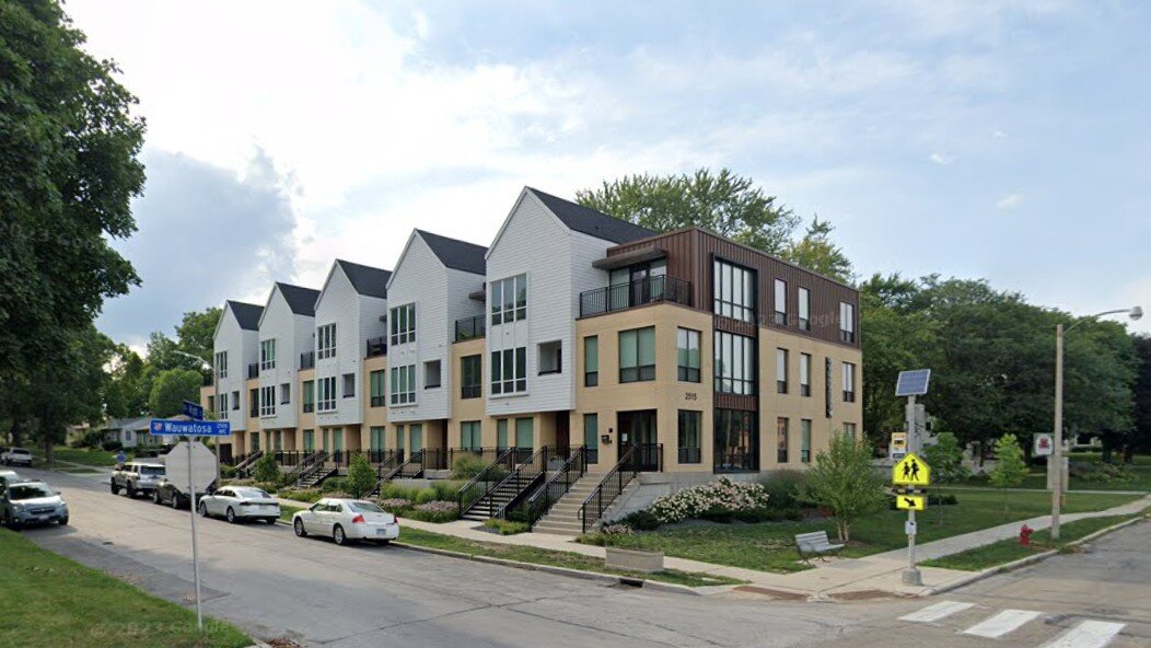 A modern row of townhouses with clean, angular designs and light-colored facades, located on a street corner. The landscaping is well-maintained, and the overall scene presents a contemporary and suburban vibe.