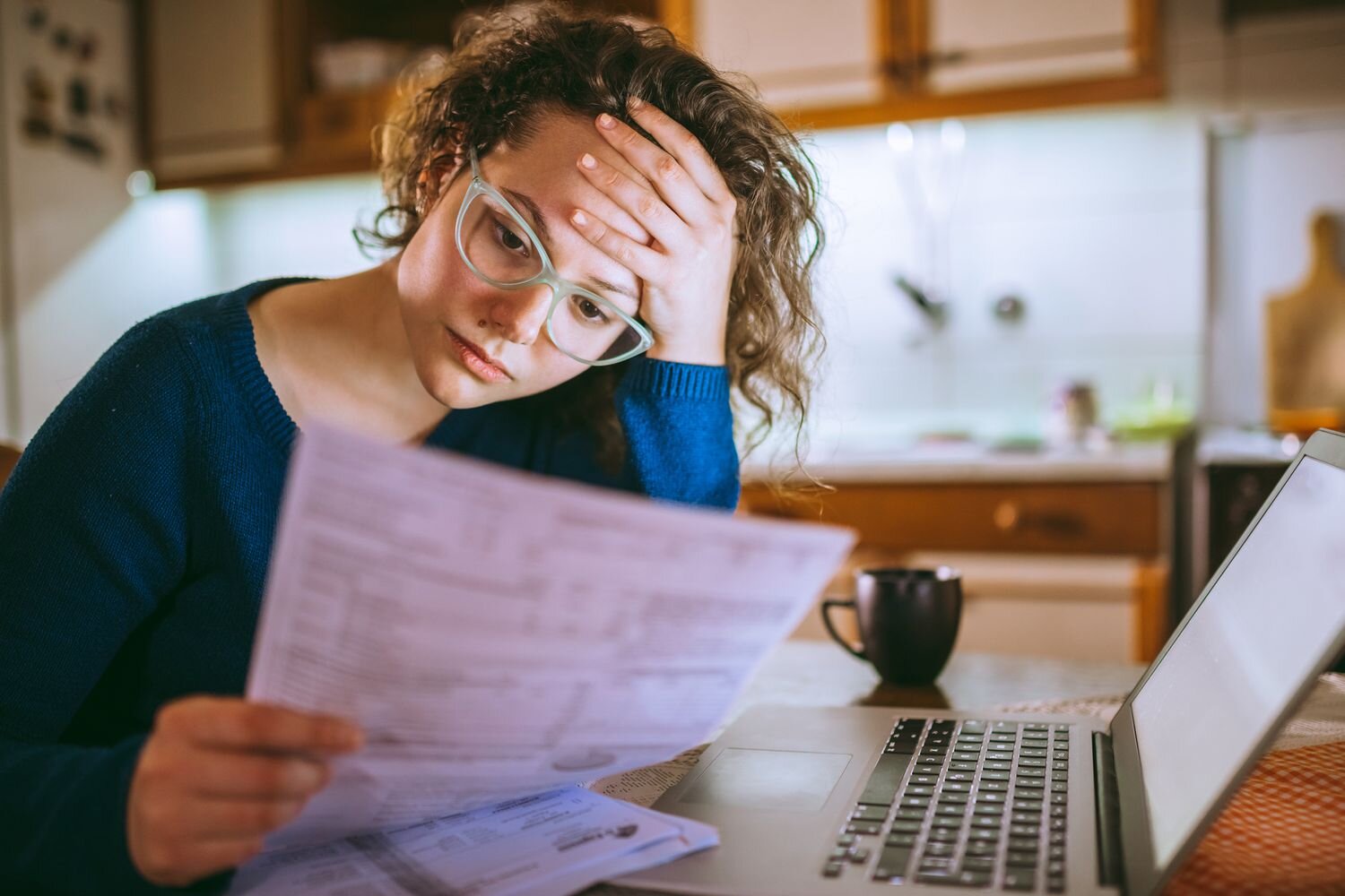 A young woman with curly hair and glasses looks worried as she reviews a financial document in her kitchen, sitting next to a laptop. She rests her hand on her forehead, depicting financial stress.
