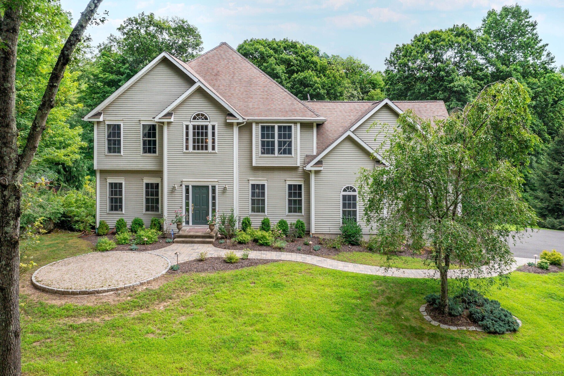 A spacious, two-story beige home with a symmetrical design, set in a lush, green, forested area. The house features a large front entrance with an arched window above the door and a gently curved walkway lined with shrubbery, leading to the main entryway.