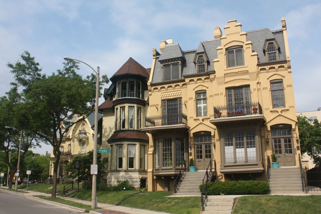 A row of historic, ornate, and yellow brick townhouses in a cityscape, showcasing intricate architectural details, large windows, and a street corner with mature trees lining the sidewalk.