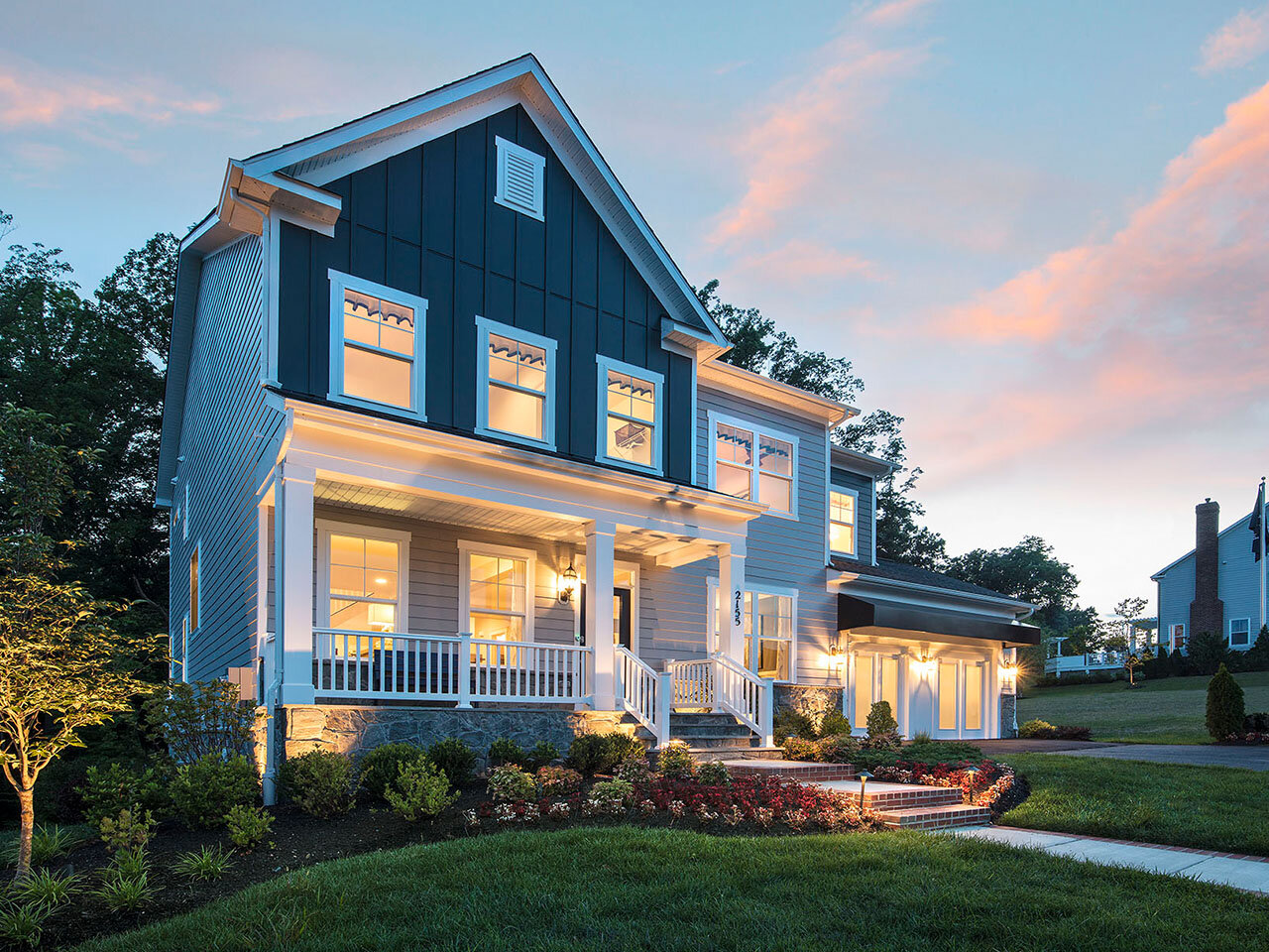 A two-story modern home at dusk with a front porch and large windows warmly illuminated from inside. The exterior combines light gray siding with a dark blue upper facade, and the landscaping includes a mix of shrubs and flowers, enhancing the welcoming ambiance.