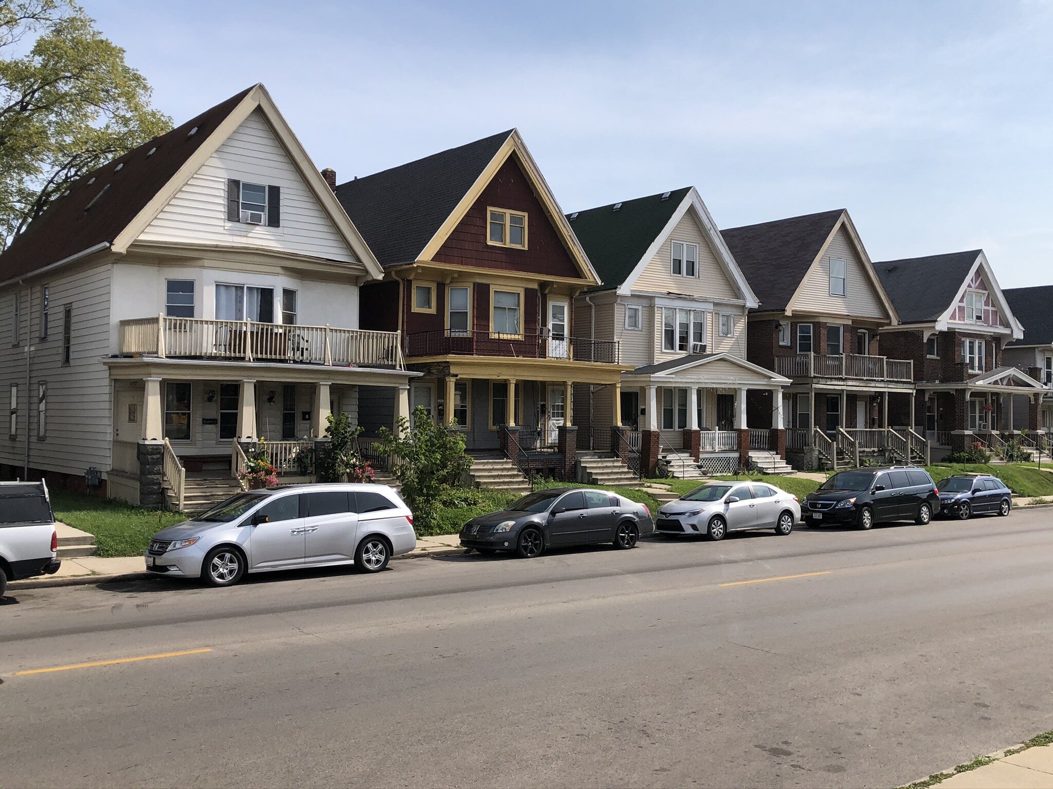 A row of traditional two-story homes along a quiet street, each with distinct architectural styles, front porches, and balconies. Parked cars line the curb, giving a suburban feel to this well-kept neighborhood scene.