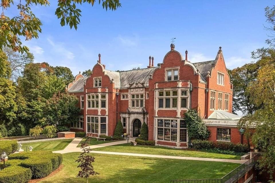 An elaborate historic mansion in red brick with intricate stone detailing and large windows. The architecture features a grand entrance with arched windows, decorative rooflines, and lush green landscaping surrounding the property, set against a blue sky.