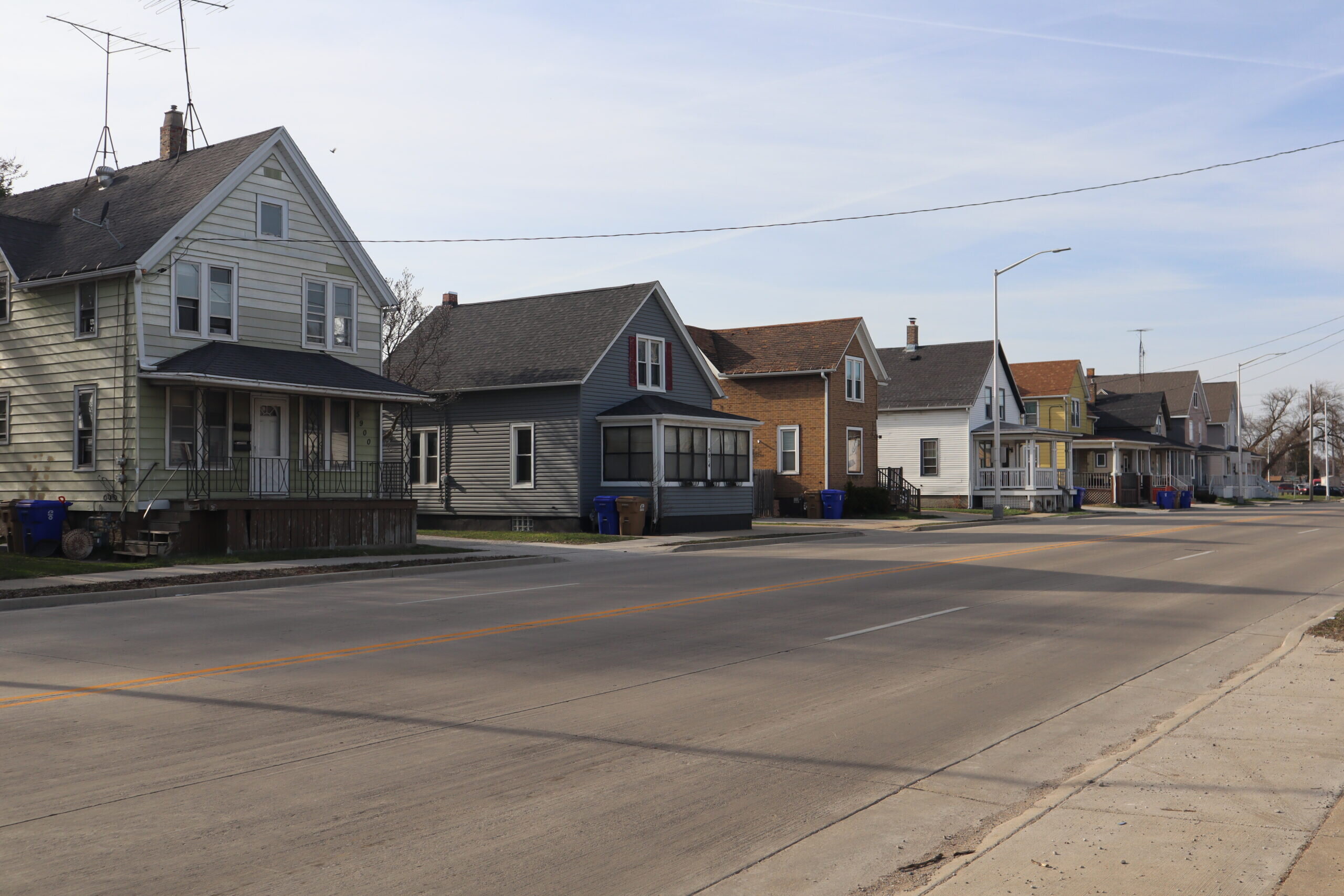 A row of small residential houses along a quiet suburban street with similar architectural styles. The houses feature different color schemes, some with porches and fenced-in front yards.
