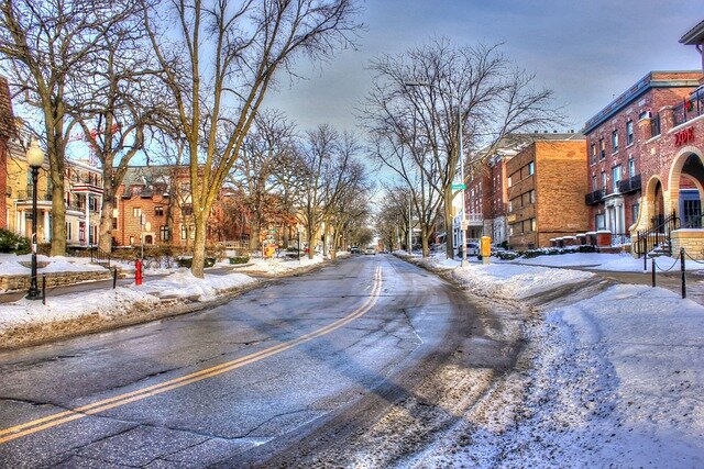 A quiet residential street in a small town, covered in light snow, with bare trees lining the road and a mix of historic and modern buildings on either side.
