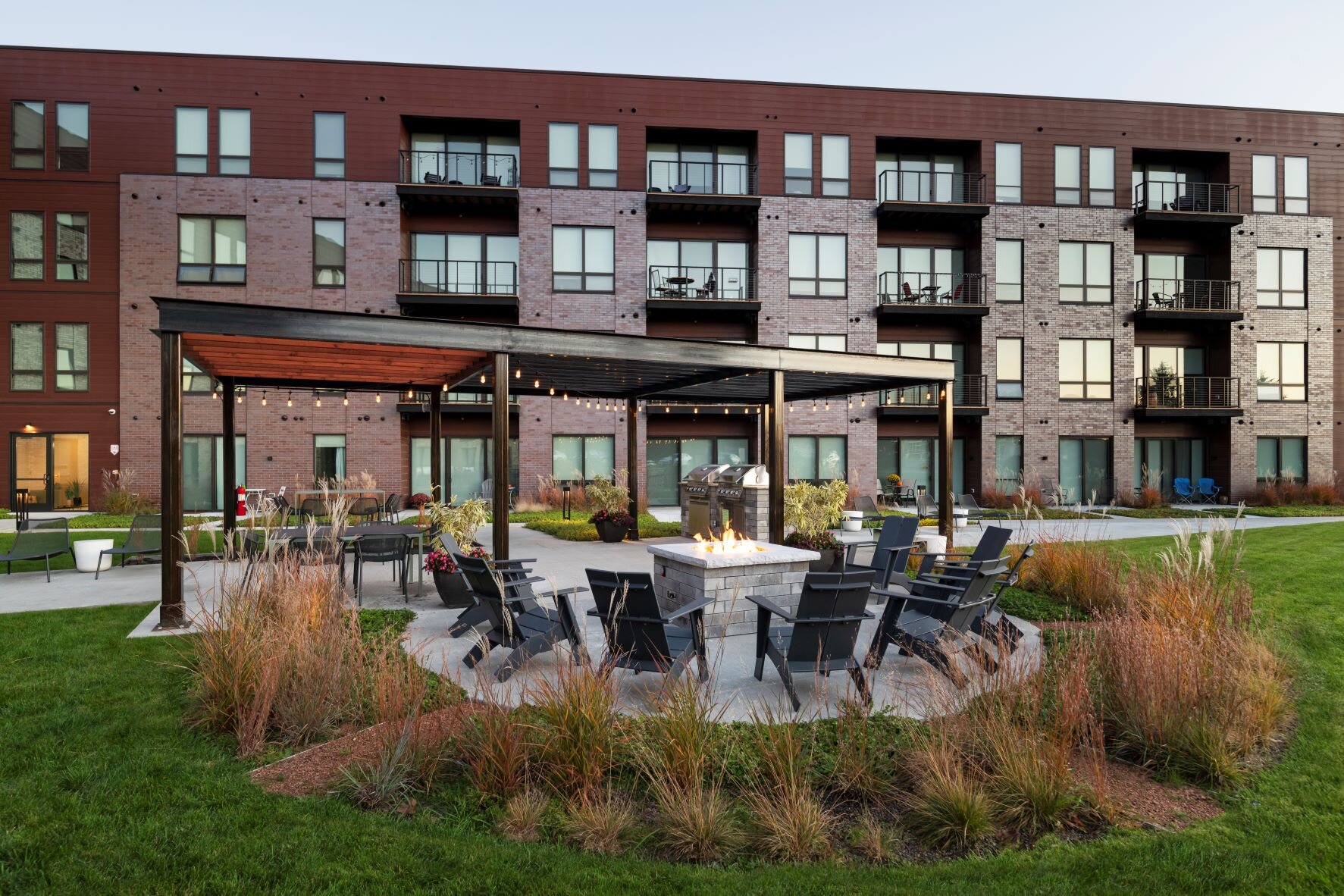 A beautifully designed outdoor courtyard of a modern brick apartment complex, featuring a cozy fire pit surrounded by black chairs, a pergola with hanging lights, and landscaped greenery, creating a welcoming atmosphere.