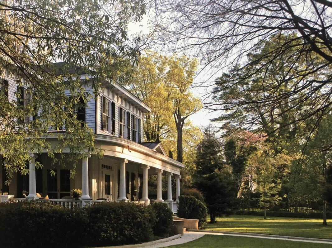 A picturesque Southern-style colonial house with a wraparound porch and classic white columns, nestled among large, mature trees. The scene is serene, with soft sunlight filtering through the leaves, casting a warm glow on the house and its lush green surroundings.